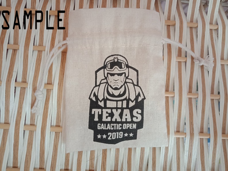 May include: A beige drawstring bag with a black graphic of a person wearing a helmet and goggles. The text "TEXAS GALACTIC OPEN 2019" is printed below the graphic. The word "SAMPLE" is printed at the top of the image.