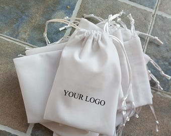 May include: A collection of white drawstring bags with the text "YOUR LOGO" printed on one of the bags. The bags are made of a soft material and have white drawstrings. The bags are arranged on a tiled surface.