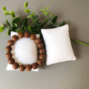 May include: A brown wooden beaded bracelet on a white pillow. The bracelet is made of round beads and has a simple design.