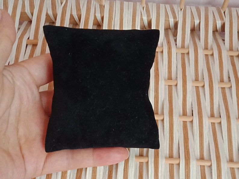 May include: A black velvet jewelry box or ring pillow. The pillow is small and square, and is sitting on a woven surface.