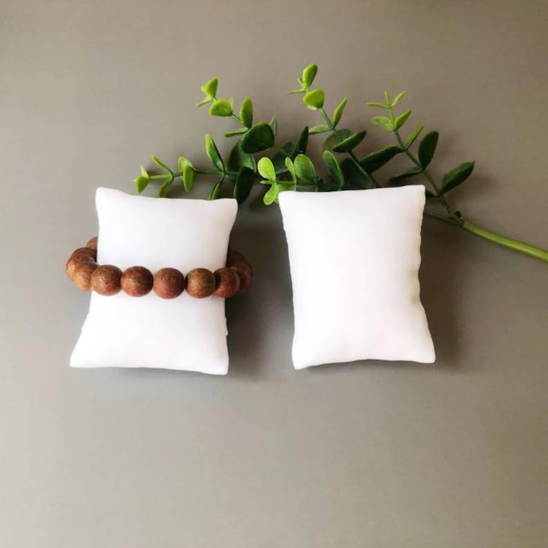 May include: Two white, square, fabric-covered display pads for jewelry. One pad has a brown wooden beaded bracelet on it.