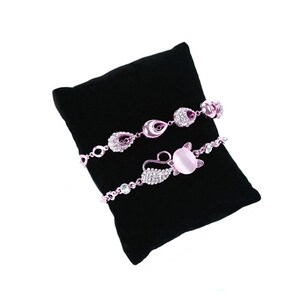 May include: Three delicate bracelets with pink and silver accents displayed on a black velvet cushion. The bracelets feature crystal embellishments and a pink stone.