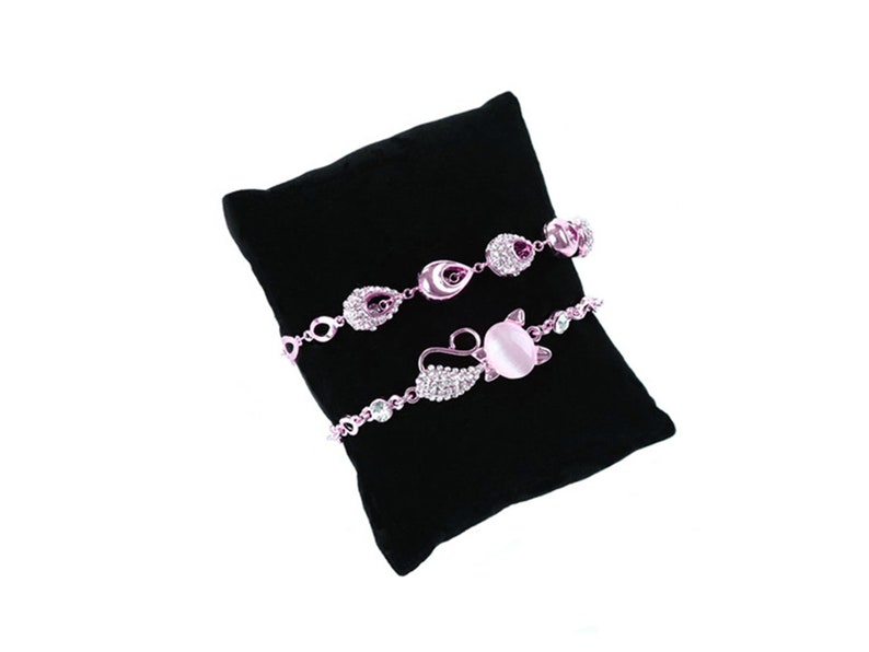 May include: Three delicate bracelets with pink and silver accents displayed on a black velvet cushion. The bracelets feature crystal embellishments and a pink stone.