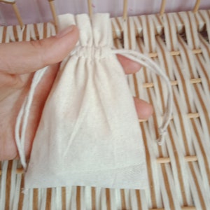 May include: A small, off-white drawstring bag made of a soft, natural fabric. The bag is held in a hand, with the drawstring partially pulled closed. The background is a woven pattern of light brown and beige.