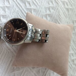 May include: A silver wristwatch with a brown face and Roman numeral markings. The watch is on a brown cushion.