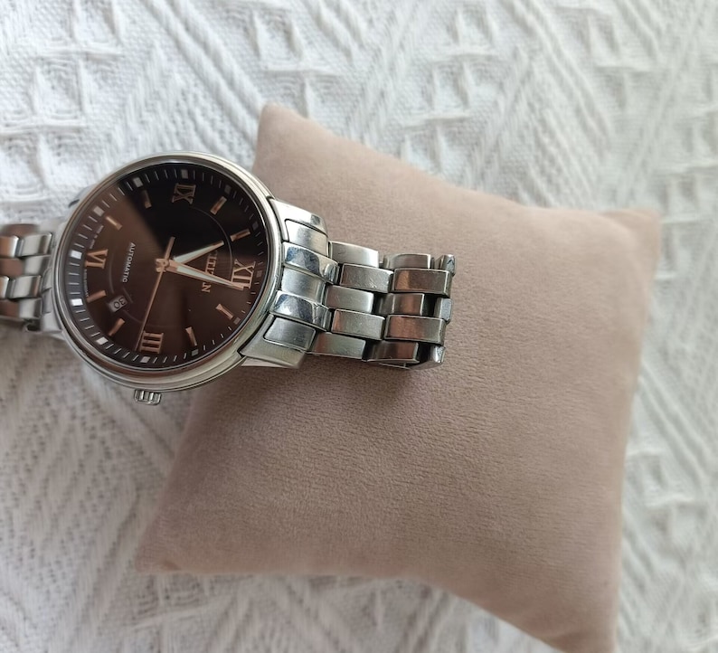 May include: A silver wristwatch with a brown face and Roman numeral markings. The watch is on a brown cushion.
