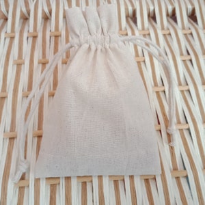 May include: A small, off-white drawstring bag made of a textured fabric. The bag is gathered at the top with a matching drawstring closure. The background is a woven pattern of light brown and white strips.