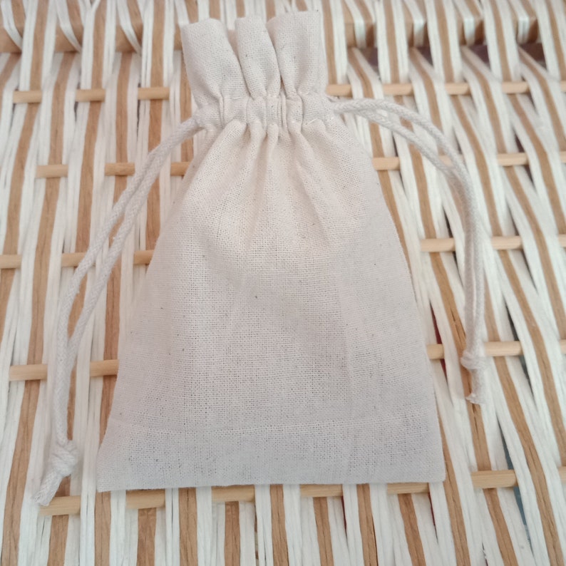 May include: A small, off-white drawstring bag made of a textured fabric. The bag is gathered at the top with a matching drawstring closure. The background is a woven pattern of light brown and white strips.
