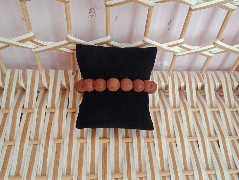 May include: A brown wooden beaded bracelet on a black velvet cushion. The bracelet is made of round beads and is displayed on a woven background.
