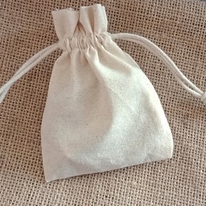 May include: A small, off-white drawstring bag made of a textured fabric. The bag is gathered at the top with a matching drawstring closure. The background is a rough, woven material, providing a natural, rustic aesthetic. The bag is likely used for small gifts or storage.