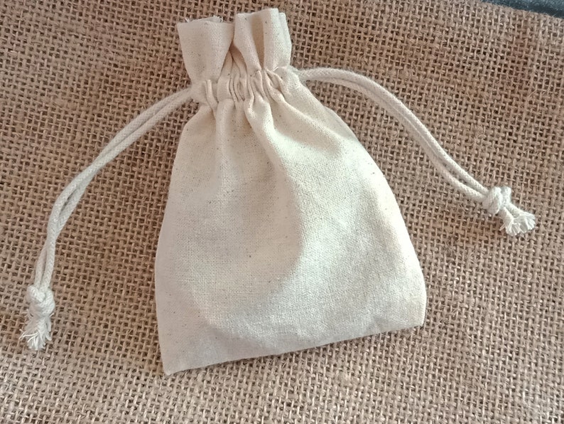 May include: A small, off-white drawstring bag made of a textured fabric. The bag is gathered at the top with a matching drawstring closure. The background is a rough, woven material, providing a natural, rustic aesthetic. The bag is likely used for small gifts or storage.