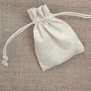 May include: A small, off-white drawstring bag made of a textured fabric. The bag is closed with a white drawstring. The background is a rough, tan fabric.