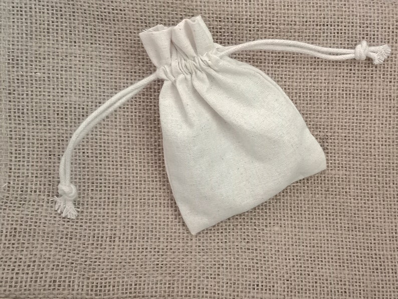 May include: A small, off-white drawstring bag made of a textured fabric. The bag is closed with a white drawstring. The background is a rough, tan fabric.