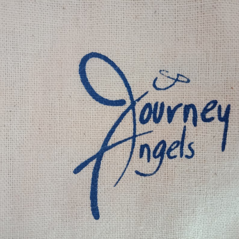 May include: Close-up of a canvas material with the words "Journey Angels" in a blue script font. The text is centered and takes up a large portion of the image. The background is a light beige color.