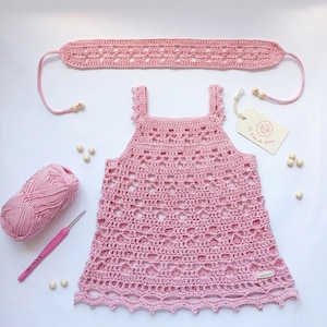 May include: A light pink crochet dress with a matching headband, a ball of matching yarn, and a pink crochet hook. The dress features an openwork pattern, a square neckline, and a small tag with a sheep illustration and the text "L'ou de Milou".