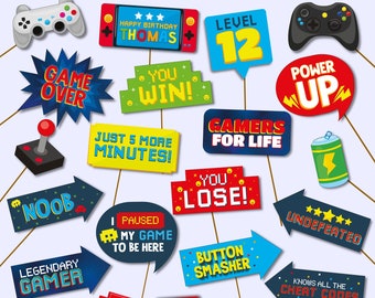 Może przedstawiać: Zestaw 17 kolorowych rekwizytów do fotobudki w temacie gier wideo. Rekwizyty obejmują kontroler gry, znak „Game Over”, znak „You Win”, znak „Level 12”, znak „Power Up”, znak „Gamers for Life”, znak „Noob”, znak „Legendary Gamer”, znak „Just 5 More Minutes”, znak „You Lose”, znak „Paused My Game to Be Here”, znak „Button Smasher”, znak „Undefeated”, znak „Knows All the Cheat Codes”, pikselowany statek kosmiczny, czerwone koło z żółtym środkiem i przenośną konsolę do gier.