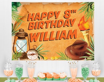 Puede incluir: Una pancarta de cumpleaños marrón y naranja con el texto "Happy 8th Birthday William". La pancarta presenta una brújula, una linterna, un sombrero y una antorcha. La pancarta se muestra sobre una mesa con cupcakes, tarros de caramelos y otras decoraciones.