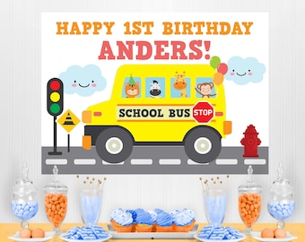 May include: A yellow school bus birthday banner with the text "HAPPY 1ST BIRTHDAY ANDERS!" features cartoon animals. The table is set with cupcakes, candies, and cookies, creating a festive party scene.