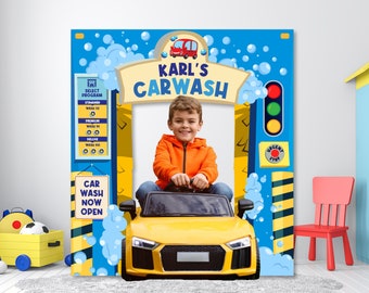 Puede incluir: Un telón de fondo de cabina de fotos de lavado de coches azul y amarillo con un niño en un coche de juguete amarillo. El telón de fondo tiene un cartel que dice "Karl's Carwash" y un menú de lavado de coches. El niño sonríe y sostiene el volante.