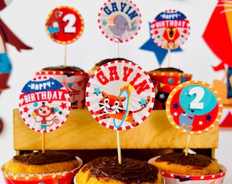 May include: A set of three cupcakes decorated with a circus theme. The cupcakes are topped with chocolate frosting and have red and white striped cupcake liners. The cupcakes are decorated with round paper toppers featuring a tiger, a monkey, and a blue fish. The toppers also have the text "Gavin" and "Happy Birthday" on them.