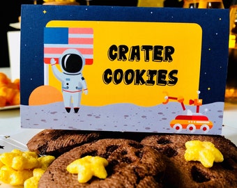 May include: Chocolate chip cookies with star-shaped cereal pieces on a white plate. A sign with the text "CRATER COOKIES" features an astronaut, the American flag, and a space rover against a yellow and blue background. The cookies are a treat for a space-themed party.