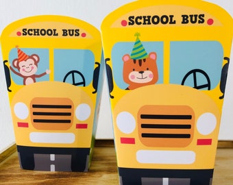 May include: Two yellow school bus-shaped party favor boxes with a monkey and a tiger wearing party hats. The boxes have the words "School Bus" printed on the front.