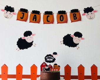 May include: A birthday cake decorated with a black sheep, a red barn, and a sign that says "Happy 1st Birthday Jacob!" The cake is on a white cake stand and in front of a brown wooden fence with green grass.
