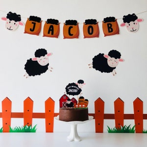 Pode incluir: Um bolo de aniversário decorado com uma ovelha preta, um celeiro vermelho e uma placa que diz "Happy 1st Birthday Jacob!" O bolo está em um suporte de bolo branco e na frente de uma cerca de madeira marrom com grama verde.