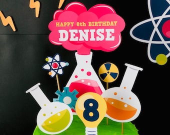 May include: A pink and white cake topper with the text "Happy 8th Birthday Denise" and a pink explosion cloud. The topper also includes a beaker, a light bulb, a gear, an atom, and a radiation symbol.