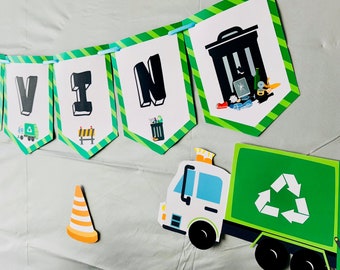 May include: A green, white, and blue recycling truck with a green recycling symbol on the side. The truck is part of a party banner that says "VIN" in green, white, and black letters. There is a green and yellow traffic cone and a red brick wall in the background.