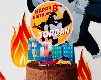 May include: A chocolate cake decorated with a King Kong themed cake topper. The topper features a cartoon King Kong smashing a city with buildings on fire. The cake topper also includes the text "Happy 8th Birthday JORDAN".