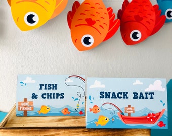 May include: Two blue and white signs with the text "Fish & Chips" and "Snack Bait". The signs have a fishing theme with cartoon fish and fishing rods. The signs are on a wooden surface with a garland of orange, red, and blue fish hanging above.