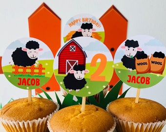 Puede incluir: Cuatro cupcakes con forros blancos y parte superior marrón. Cada cupcake tiene una parte superior redonda de papel de colores con una oveja negra y el texto "¡Feliz cumpleaños!" y "Jacob". Una parte superior tiene un granero rojo y el número "2" en ella.