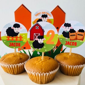 Pode incluir: Quatro cupcakes com forros brancos e topo marrom. Cada cupcake tem um topo redondo de papel colorido com uma ovelha preta e o texto "Feliz aniversário!" e "Jacob". Um topo tem um celeiro vermelho e o número "2" nele.