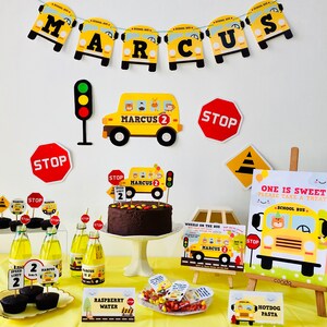 A birthday party table setting with a yellow school bus theme. The table is decorated with yellow school buses, stop signs, and a chocolate cake with a school bus topper. There are also cupcakes, sweets, and drinks on the table. The table is covered in a yellow tablecloth. The text on the cake says "Marcus 2".