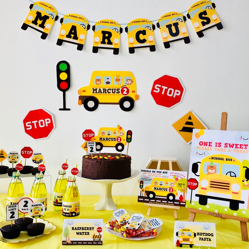 A birthday party table setting with a yellow school bus theme. The table is decorated with yellow school buses, stop signs, and a chocolate cake with a school bus topper. There are also cupcakes, sweets, and drinks on the table. The table is covered in a yellow tablecloth. The text on the cake says "Marcus 2".