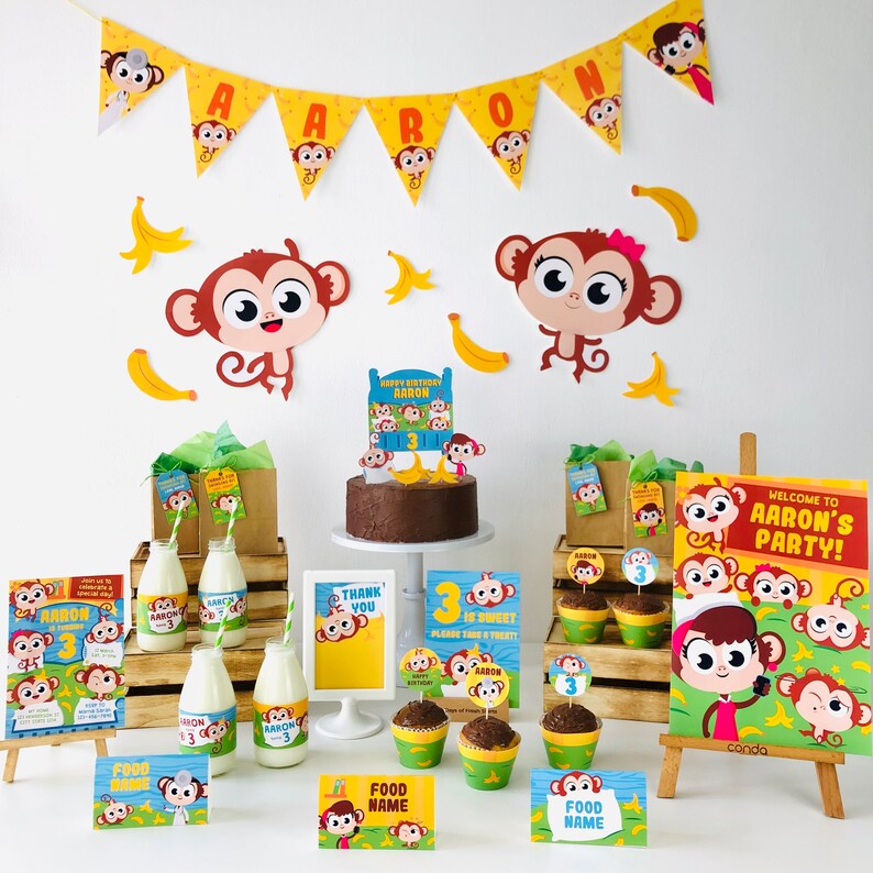 May include: A monkey-themed birthday party setup with a chocolate cake, cupcakes, milk bottles, and decorations. The decorations include a banner with the name "Aaron", a welcome sign, and food labels. The cake is decorated with a monkey-shaped topper and a number "3" candle.