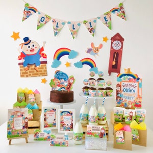 May include: A festive birthday party setup with a banner that reads "OLLIE". Includes a cake, cupcakes, milk bottles, and various decorations featuring cartoon characters, rainbows, and stars. The theme is a child's first birthday.