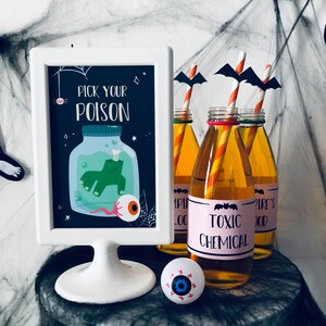 May include: A white sign with black lettering that reads "Pick Your Poison" with a cartoon illustration of a jar with a green hand inside and a red eyeball. There are four glass bottles with orange and white striped straws and black bat cutouts. The bottles have labels that read "Vampire Blood", "Toxic Chemical", and "Vampire's Blood".