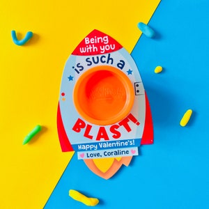 May include: A Valentine's Day card shaped like a rocket, with the text "Being with you is such a BLAST! Happy Valentine's! Love, Coraline." An orange Play-Doh container is in the center. Colorful Play-Doh pieces are scattered around the card on a yellow and blue background.