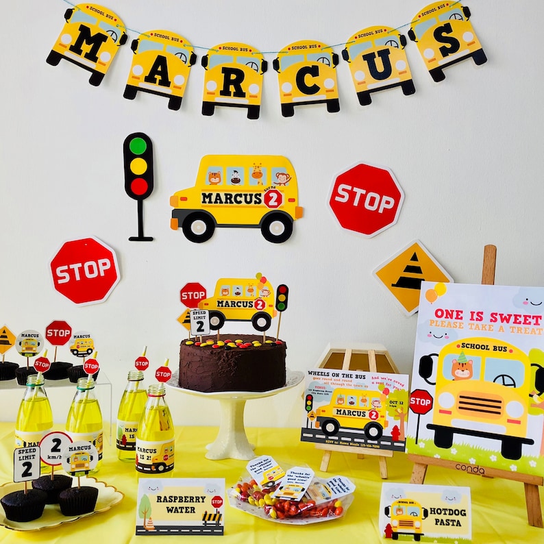 A birthday party table setting with a yellow school bus theme. The table is decorated with yellow school buses, stop signs, and a chocolate cake with a school bus topper. There are also cupcakes, sweets, and drinks on the table. The table is set on a yellow tablecloth. The text on the cake says "Marcus 2".