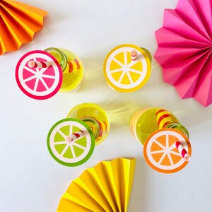 May include: Four clear glass bottles with colorful paper lids and straws. The lids are decorated with images of lemons and limes. The bottles are sitting on a white surface with colorful paper fans.