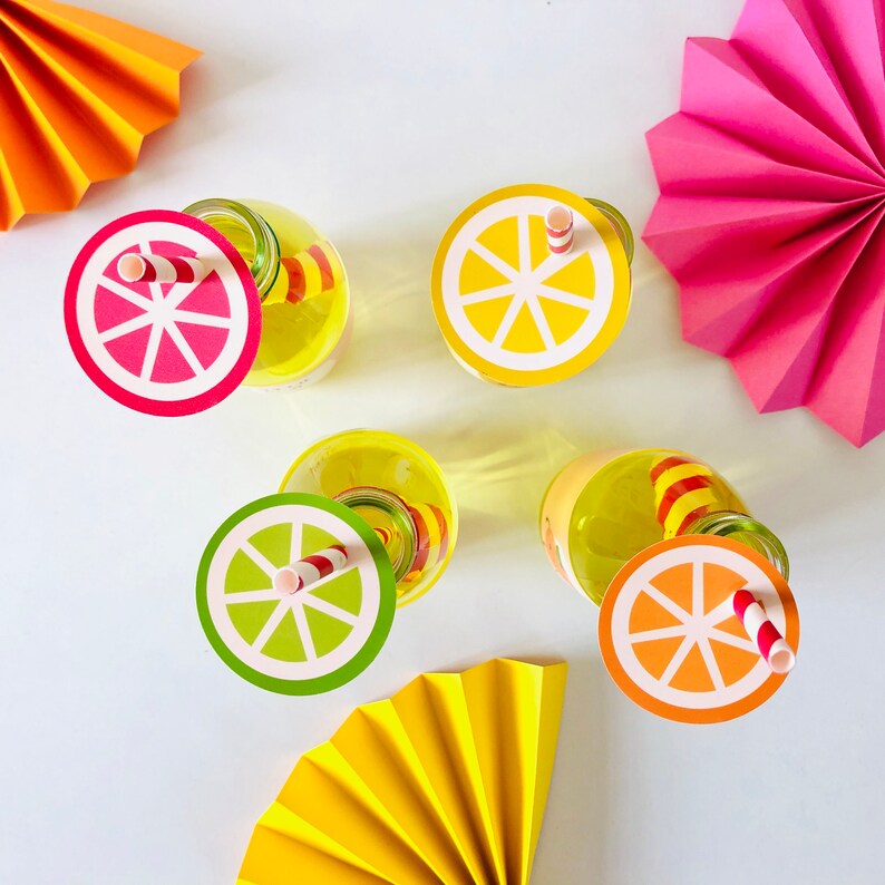 May include: Four clear glass bottles with colorful paper lids and straws. The lids are decorated with images of lemons and limes. The bottles are sitting on a white surface with colorful paper fans.