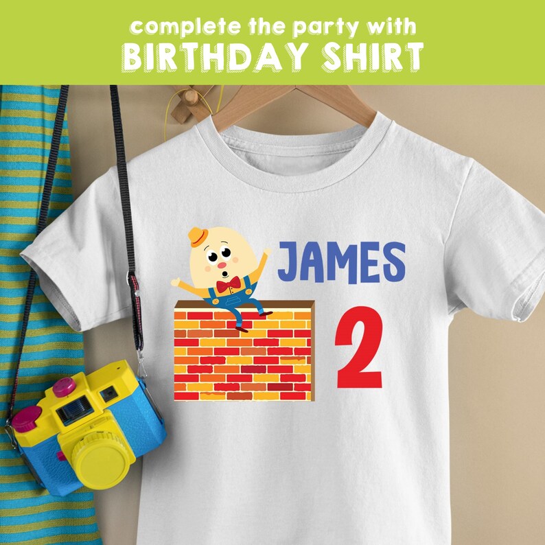 Puede incluir: Camiseta blanca con un personaje de dibujos animados de un huevo amarillo con una pajarita roja y un mono azul sentado en un muro de ladrillos. El texto "JAMES 2" est&aacute; impreso en la camiseta.