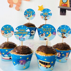 May include: Blue and yellow cupcake wrappers with a star and moon theme. The wrappers feature a variety of characters, including a sheep, a spider, a cat, and a bus. The cupcakes are topped with chocolate frosting and decorated with round toppers that say "Ollie", "One", and "Happy Birthday!"