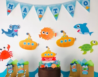 May include: A blue and white banner with the name Matteo on it. There are several decorations on a white wall, including a yellow submarine, a blue shark, a green turtle, a red crab, and an orange pufferfish. A chocolate cake with a blue shark, a green turtle, and an orange pufferfish on top is on a white cake stand. The cake is decorated with a red ribbon that says "Happy 4th Birthday Matteo". There are also several brown paper bags with blue and white tags that say "Thanks".
