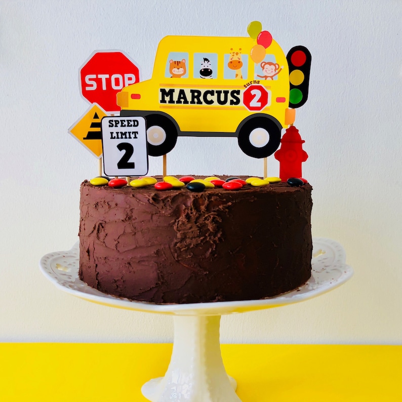A Wheels on the Bus themed birthday cake decorated with red and black sweets and topped with a yellow school bus cake topper with the name "Marcus 2" on it. The bus has a stop sign, a speed limit sign, and a traffic light.