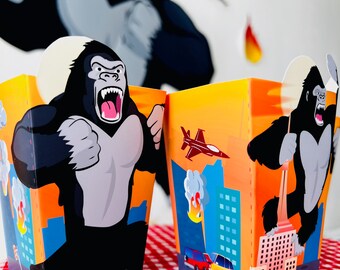 May include: Two yellow and orange party favor boxes with a gorilla graphic. One box features a gorilla climbing a building and the other features a gorilla holding a plane. Both boxes have a black and white gorilla graphic.