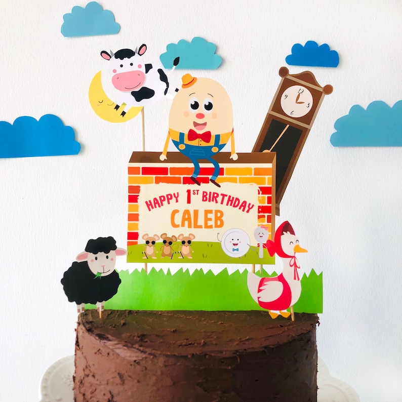 K&ouml;nnte beinhalten: Ein Schokoladenkuchen mit einem Schild Happy 1st Birthday Caleb, einer roten Backsteinmauer, einem gr&uuml;nen Grasfeld, einer Kuh mit einem Mond, einer Uhr, einem schwarzen Schaf, einem wei&szlig;en Ei mit einer roten Schleife und einer rot-wei&szlig;en Gans.