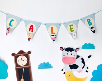 May include: A blue and white banner with the letters CALEB in colorful letters. The banner is strung up on a white wall with a clock, clouds, and a cow with a moon under it.